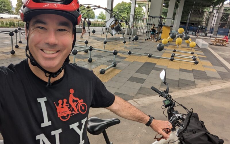 Patrick Weir wearing a red helmet on a cargo bike in New York City near a playground