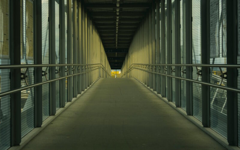 Covered pedestrian bridge over railway tracks, with light at the far end — symbolizing quiet progress and crossing into something new.